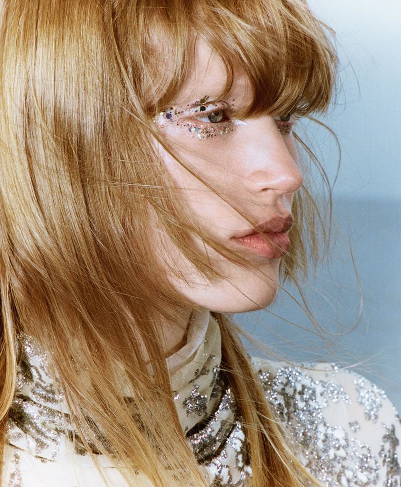 Close-up portrait of a young woman with blond hair and silver glitter makeup around her eye, posing against a muted blue background.