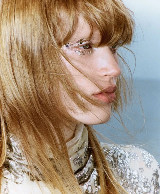 Close-up portrait of a young woman with blond hair and silver glitter makeup around her eye, posing against a muted blue background.