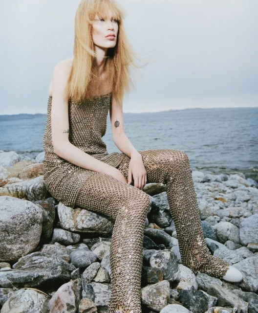 A blonde woman with pale skin poses on rocks near the sea, wearing a gold mesh top and pants, with a cloudy sky background.