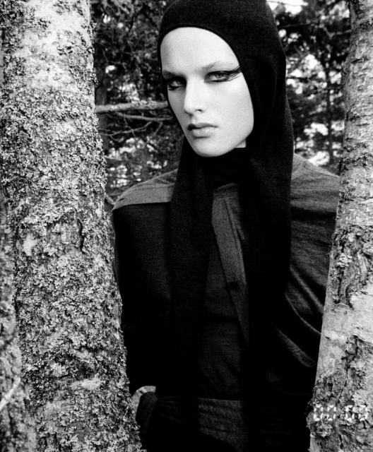 Black and white portrait of a woman in a black hooded garment, standing outdoors between two trees in a wooded area.
