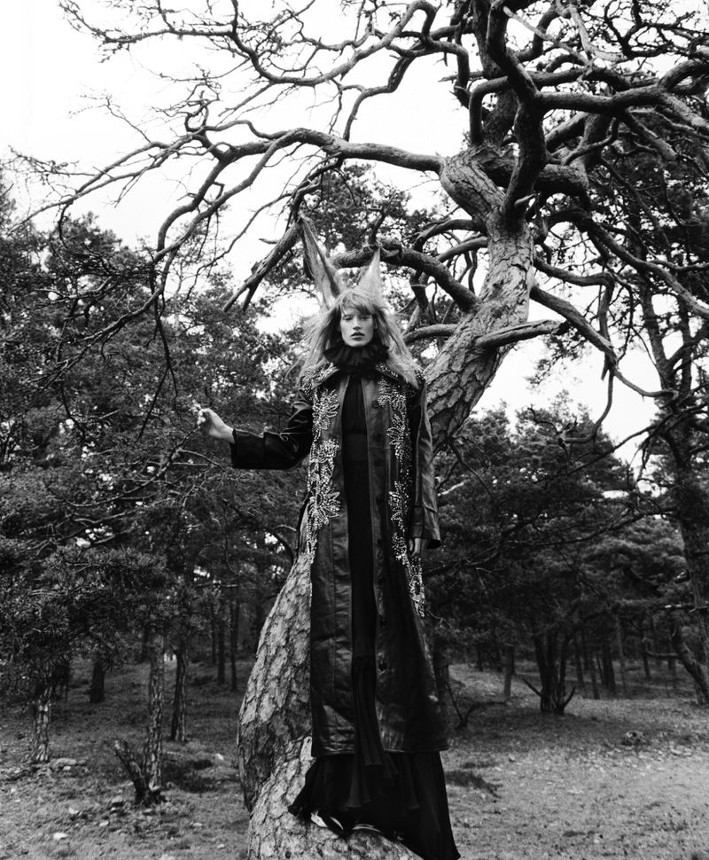 Black and white portrait of a woman in an ornate coat with a horned headpiece posing next to a tree in a forest landscape.