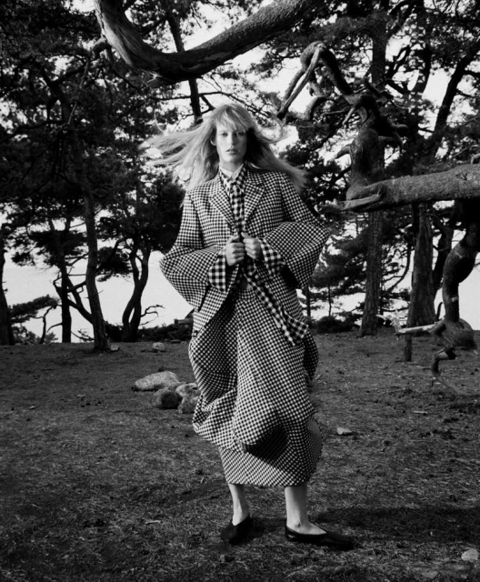 A woman in a patterned jacket and dress stands in a black and white photograph outdoors near trees on a grassy field.