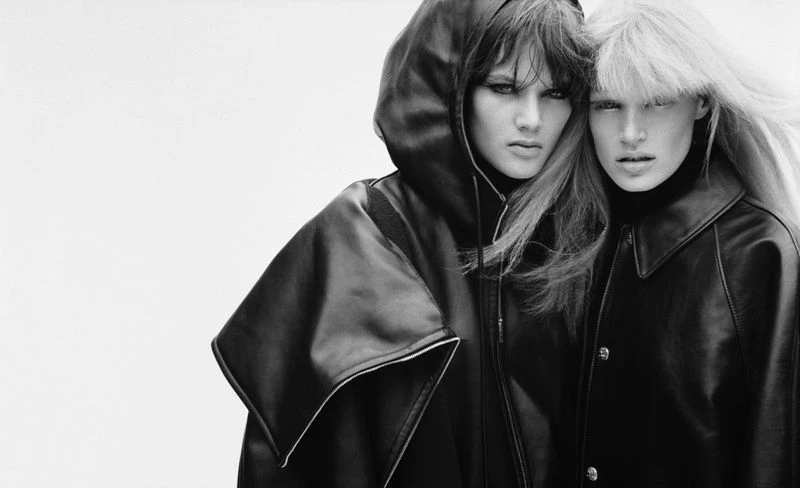 Two fashion models in black turtlenecks and leather jackets with a white background. One model has a hooded leather jacket.