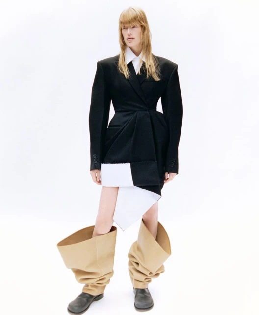 A woman poses in a black oversized blazer with a white shirt and skirt, paired with exaggerated beige boots against a white backdrop.