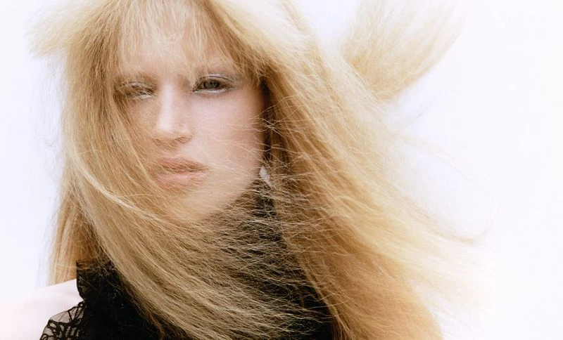 Close-up portrait of an adult female with teased blonde hair and lace garment against a bright white background in a studio setting.
