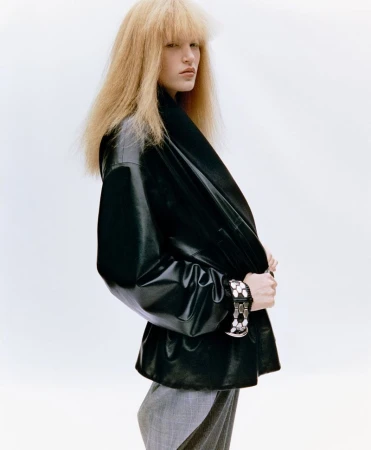 Model with voluminous blonde hair wearing an oversized black leather jacket and grey trousers against a bright white background.