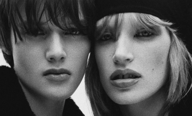 Close-up black and white portrait of a young adult with dark hair and a fair-skinned woman wearing a black beret. Editorial fashion.