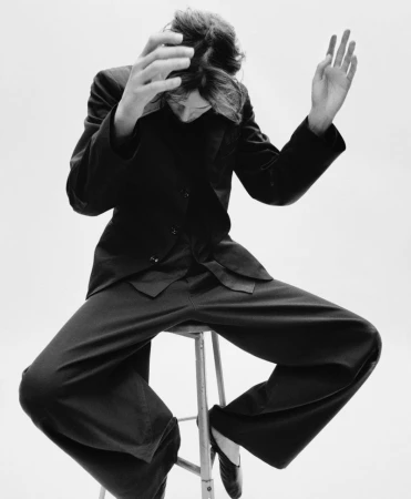 Monochrome fashion portrait of a person in a black suit sitting on a stool with hands raised against a white background in studio lighting.
