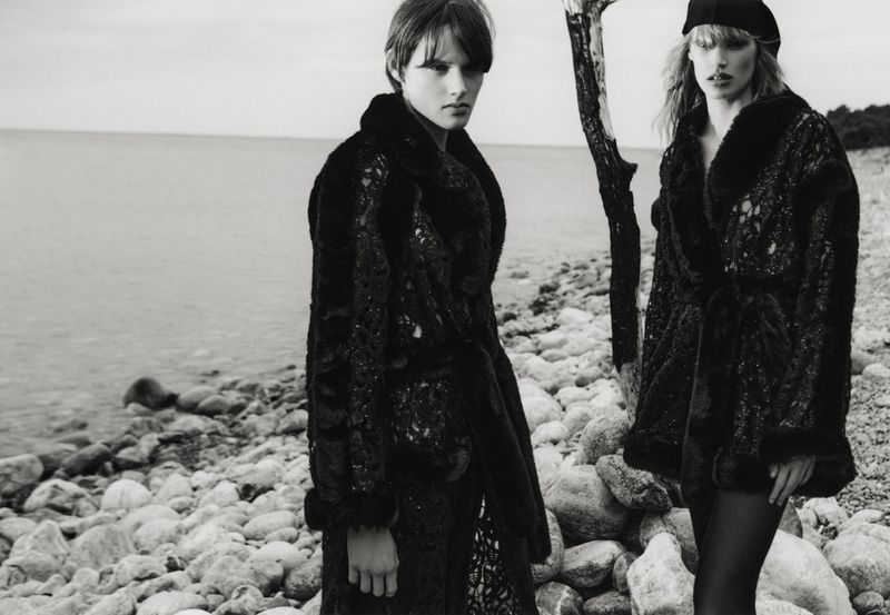 Two women in dark, textured coats pose on a rocky shoreline next to the water, wearing hats and facing the camera in a black and white fashion shot.