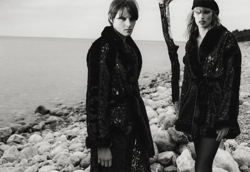 Two women in dark, textured coats pose on a rocky shoreline next to the water, wearing hats and facing the camera in a black and white fashion shot.