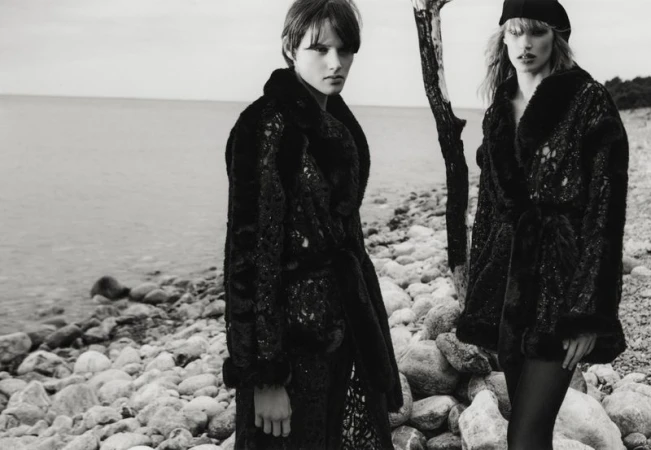 Two women in dark, textured coats pose on a rocky shoreline next to the water, wearing hats and facing the camera in a black and white fashion shot.
