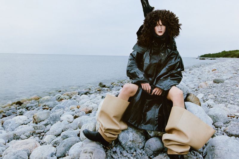 A fashion portrait shows an adult woman in an oversized black coat and tan boots, seated on a rocky beach next to calm water.