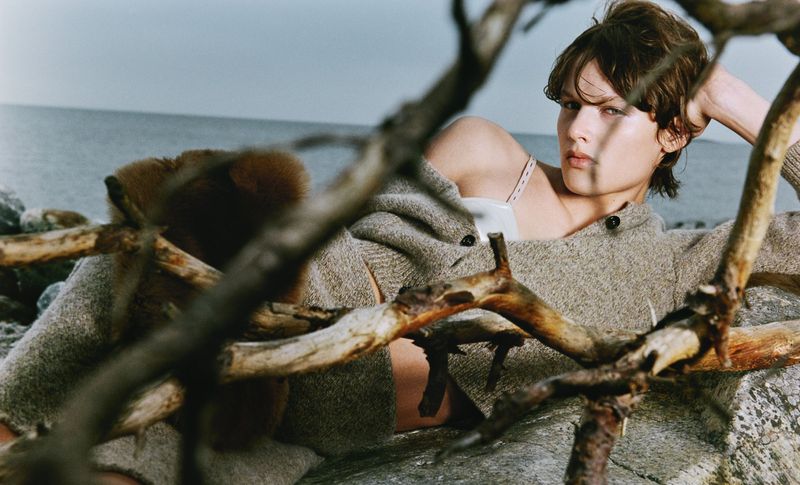 A woman in casual fashion leans back on a rocky coast, seen through tangled branches, with a sea backdrop under soft, natural light.