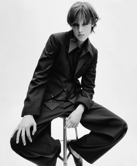 Monochromatic fashion portrait of a young adult posing on a stool wearing a dark blazer, button-down shirt and flared pants in studio.