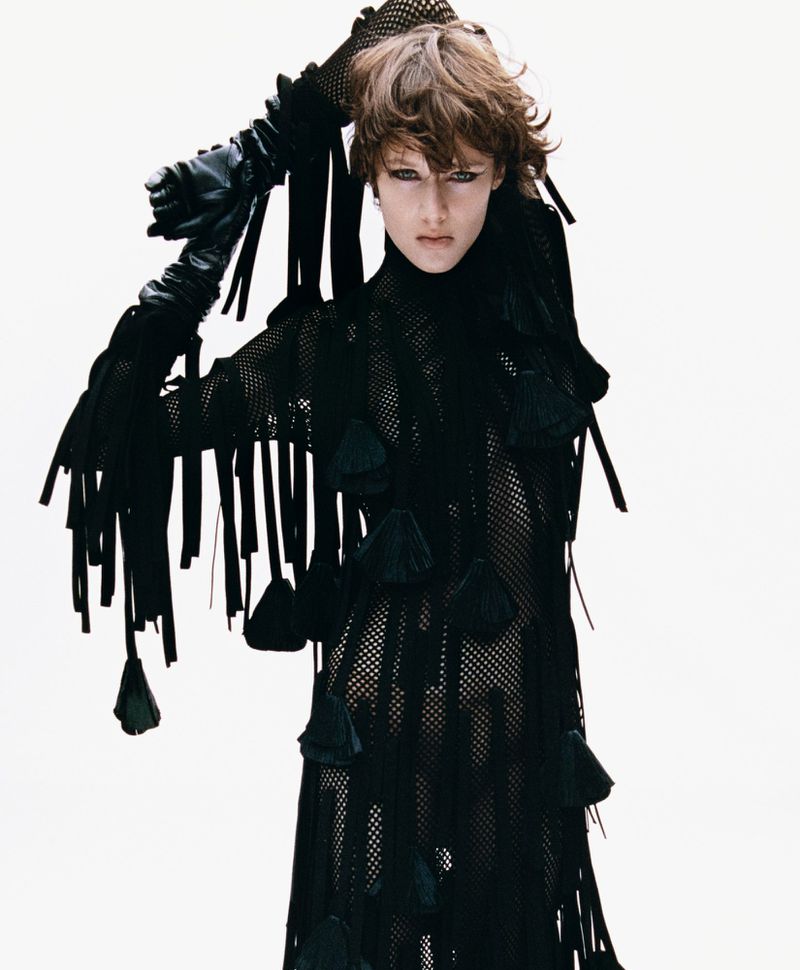 A woman with tousled brunette hair poses in a black, net-like dress with decorative tassels and black leather gloves against a white backdrop.
