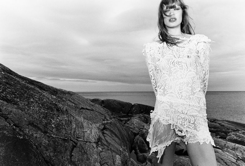 A woman in a white lace dress stands on a rocky beach in a black and white fashion shot, with the ocean and cloudy sky in the background.