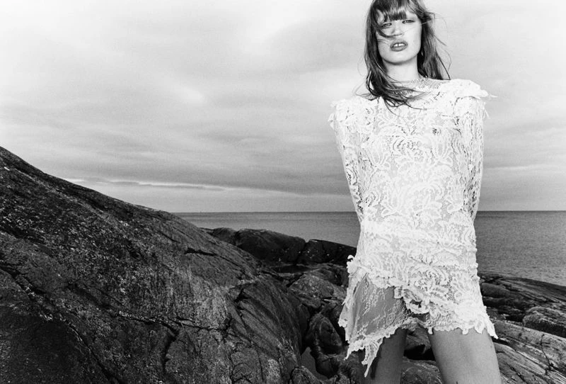 A woman in a white lace dress stands on a rocky beach in a black and white fashion shot, with the ocean and cloudy sky in the background.