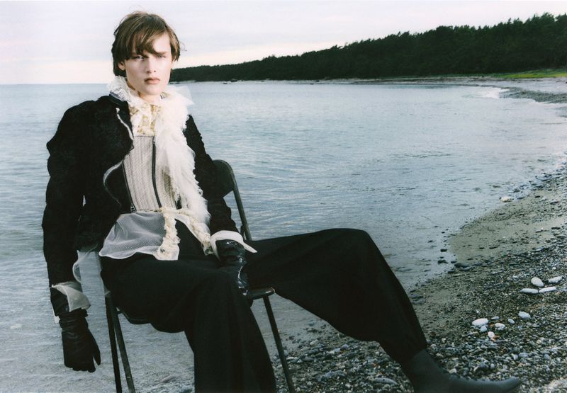 Fashionable individual sits on a chair at the beach with the ocean and trees in the background, wearing a Victorian-style top and black outfit.