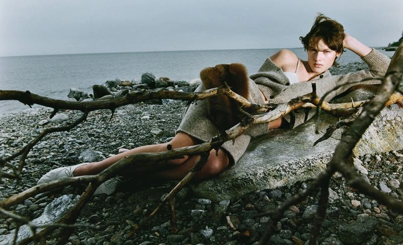 Woman lying on a rock at a rocky beach, wearing a cardigan, with the ocean in the background and branches in the foreground.