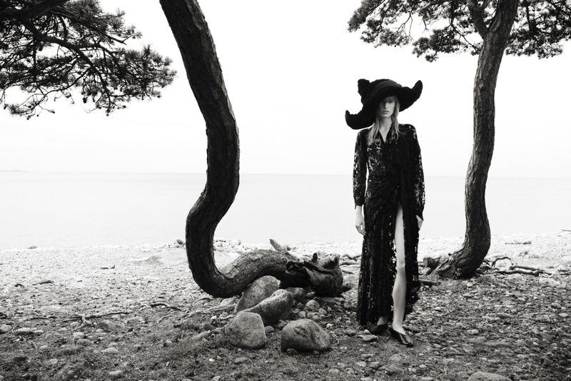 Monochrome image of a female fashion model in an outdoor setting wearing a large hat and long black dress, posing near trees and water.
