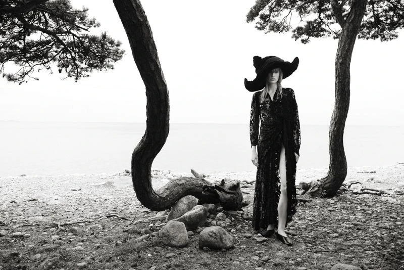 Monochrome image of a female fashion model in an outdoor setting wearing a large hat and long black dress, posing near trees and water.