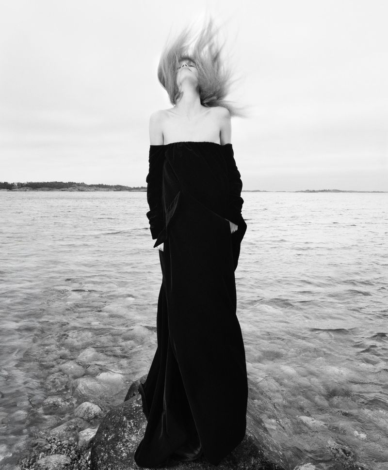 Black and white portrait of woman tossing hair on rocky shore with ocean backdrop. Moody, dramatic image, full-length shot.