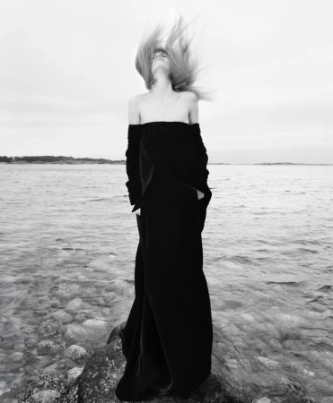 Black and white portrait of woman tossing hair on rocky shore with ocean backdrop. Moody, dramatic image, full-length shot.
