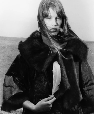 Monochrome portrait of a model with bangs wearing a dark fur-trimmed coat and holding a single white feather outdoors.