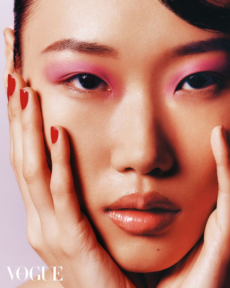 Close-up of an Asian woman's face with pink eyeshadow and red nails, hands near cheeks, glossy lips, soft lighting, and Vogue logo.