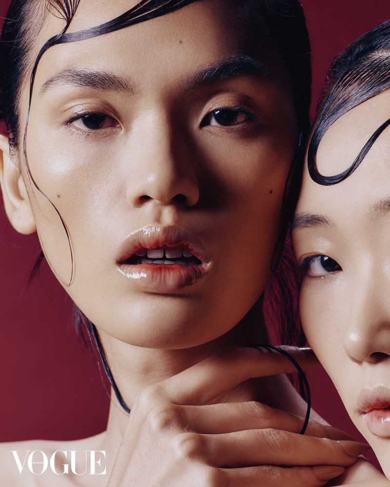 Close-up of two Asian women with glossy skin and stylized wet hair against a burgundy background, showcasing beauty and fashion elements.