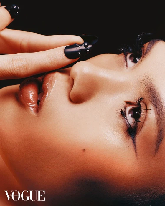 Close-up beauty portrait of an East Asian woman with glossy lips, winged eyeliner, and sculpted black nail art against a dark background.