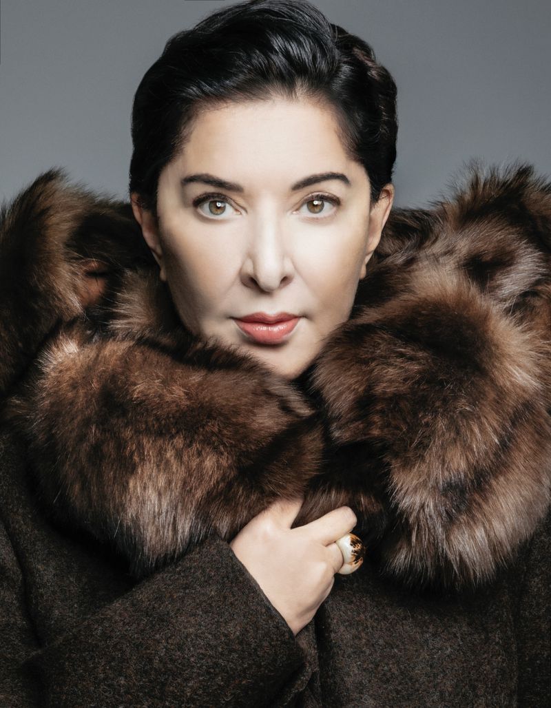 Portrait of artist Marina Abramović wearing a dark coat with a voluminous brown fur collar against a grey studio backdrop.