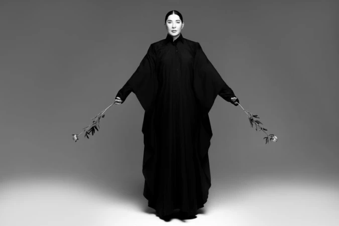 Monochrome portrait of a woman in a draped black garment, holding roses in each hand, set against a minimalist gray background.