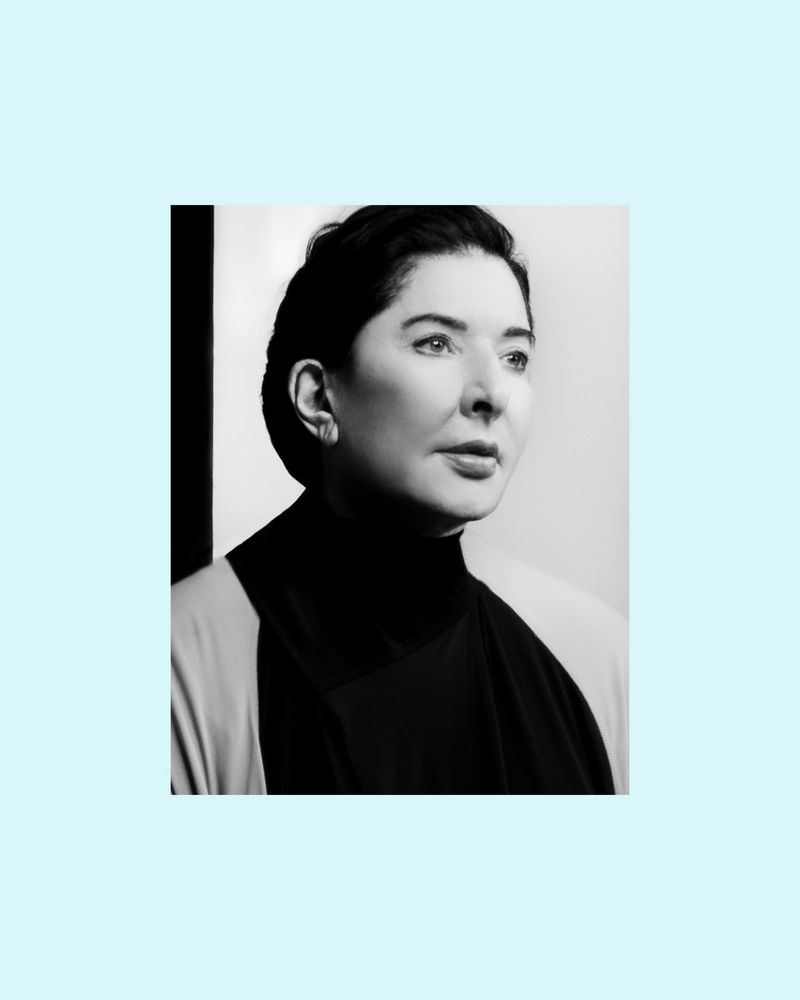 Monochrome portrait of artist Marina Abramović wearing a dark high-neck garment against a minimalist studio background.
