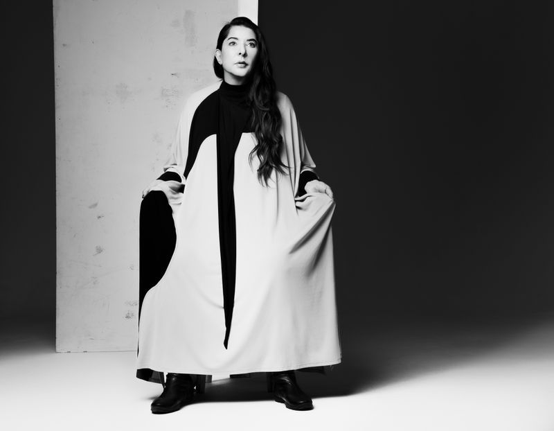 Monochrome portrait of a woman with long dark hair wearing a long, light-colored coat with black details against a grey backdrop.