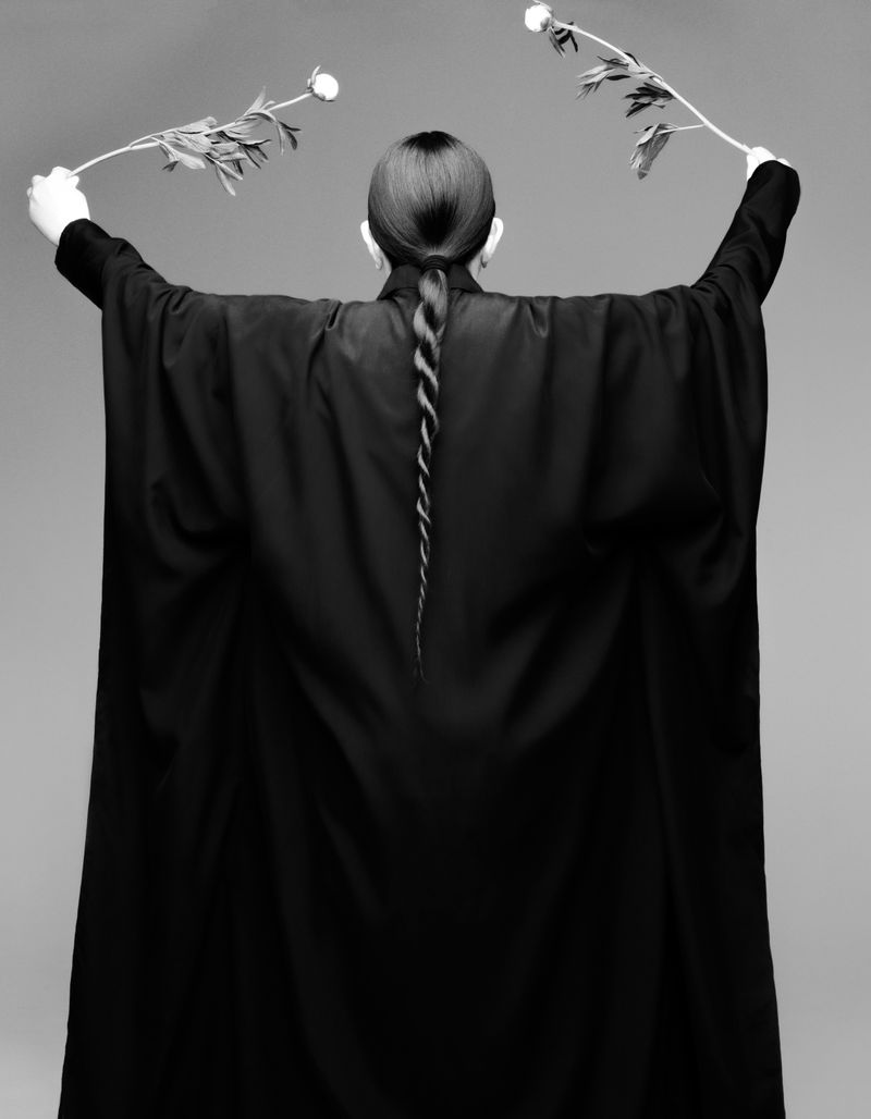 Person in black robe stands with back to camera holding floral branches, artistic black and white fashion portrait on grey background.