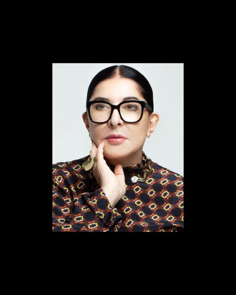 Close-up of a mature adult woman with hand near her face, wearing glasses, and a patterned top, against a bright studio backdrop.