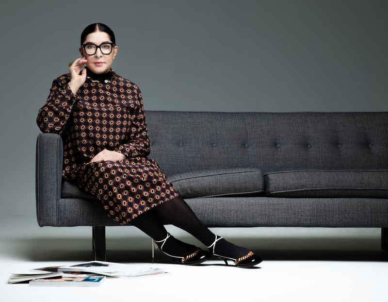 A woman with black hair and large glasses sits on a gray couch in a patterned dress in a studio, in a medium studio shot.