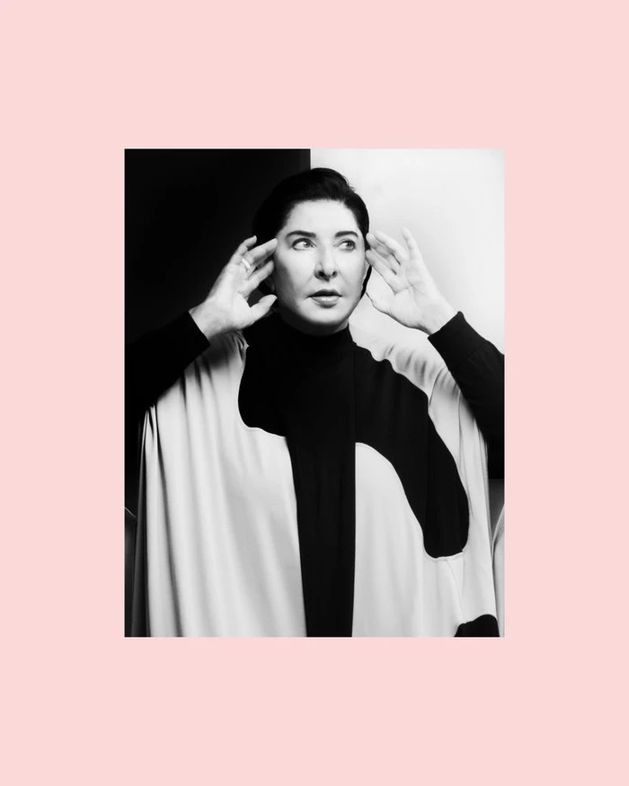 Portrait of Marina Abramović holding hands to her temples against a split black and white backdrop framed in pale pink.