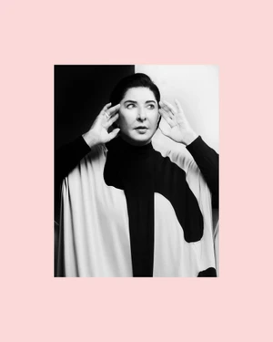Portrait of Marina Abramović holding hands to her temples against a split black and white backdrop framed in pale pink.