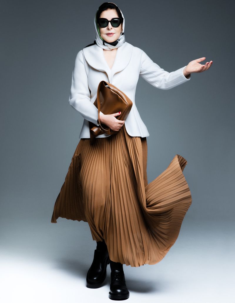 Marina Abramović wearing a white jacket, brown pleated skirt, headscarf, and sunglasses while holding a brown leather bag against a grey background
