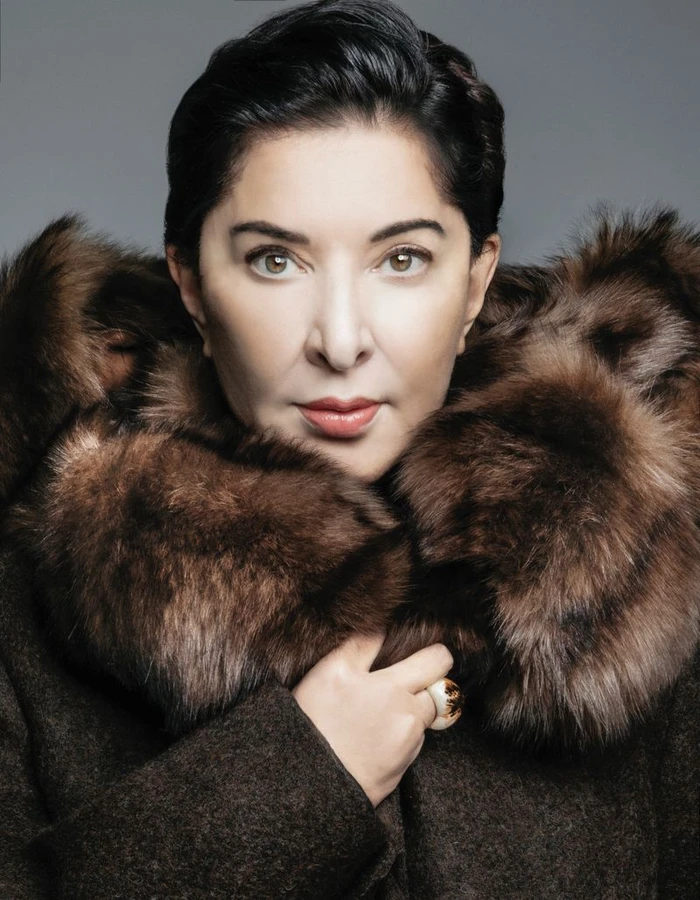 Portrait of artist Marina Abramović wearing a dark coat with a voluminous brown fur collar against a grey studio backdrop.