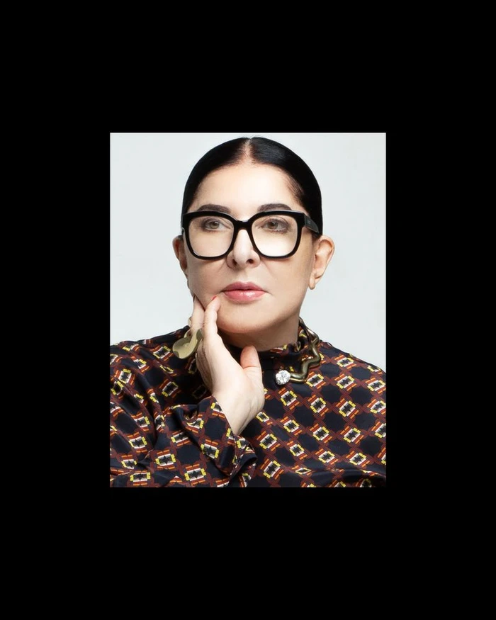 Portrait of Marina Abramovic wearing black-rimmed glasses and a patterned blouse, with hand raised to chin