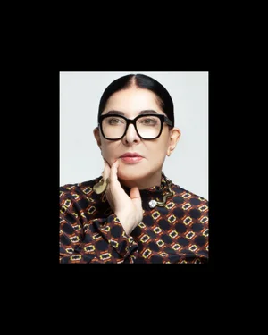Portrait of Marina Abramovic wearing black-rimmed glasses and a patterned blouse, with hand raised to chin