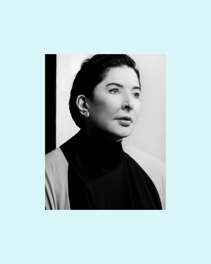 Monochrome portrait of artist Marina Abramović wearing a dark high-neck garment against a minimalist studio background.
