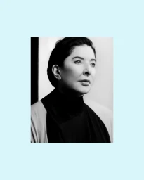 Monochrome portrait of artist Marina Abramović wearing a dark high-neck garment against a minimalist studio background.