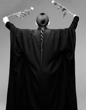 Monochrome portrait from behind of Marina Abramovic holding two flowering stems against a seamless grey backdrop.