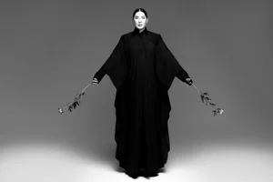 Artist Marina Abramović stands in a voluminous full-length black robe holding long-stem flowers against a grey studio backdrop.