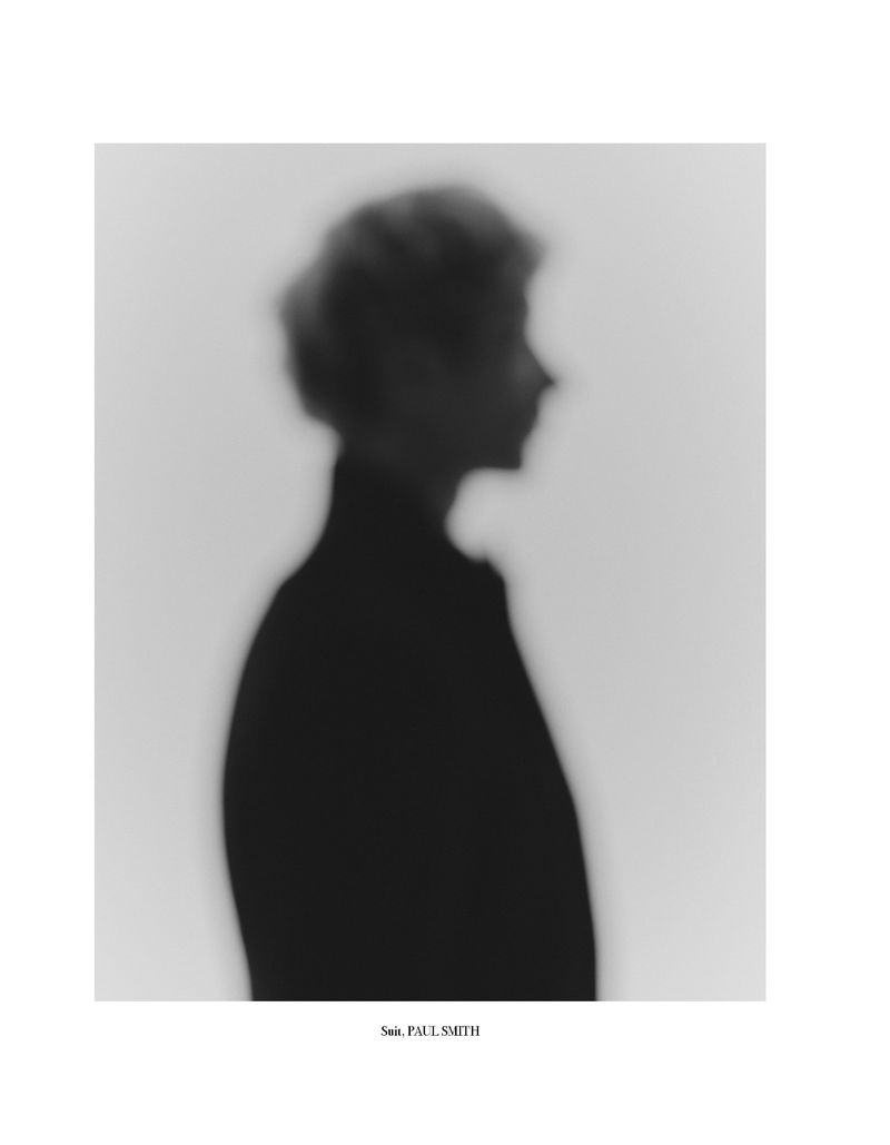 Monochromatic profile portrait of an adult person with short, curly hair in a blurred out, black and white, indoor composition.
