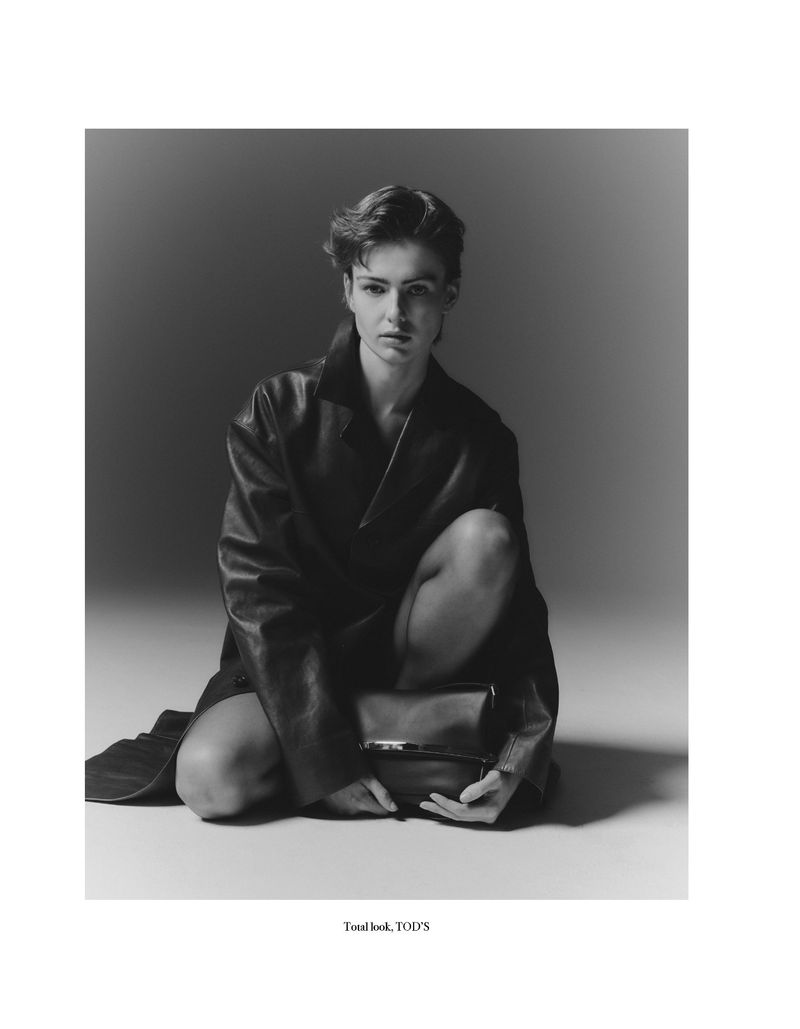 Monochrome fashion shot of a woman in a dark coat, kneeling and holding a dark bag, set against a plain grey background in a photo studio.